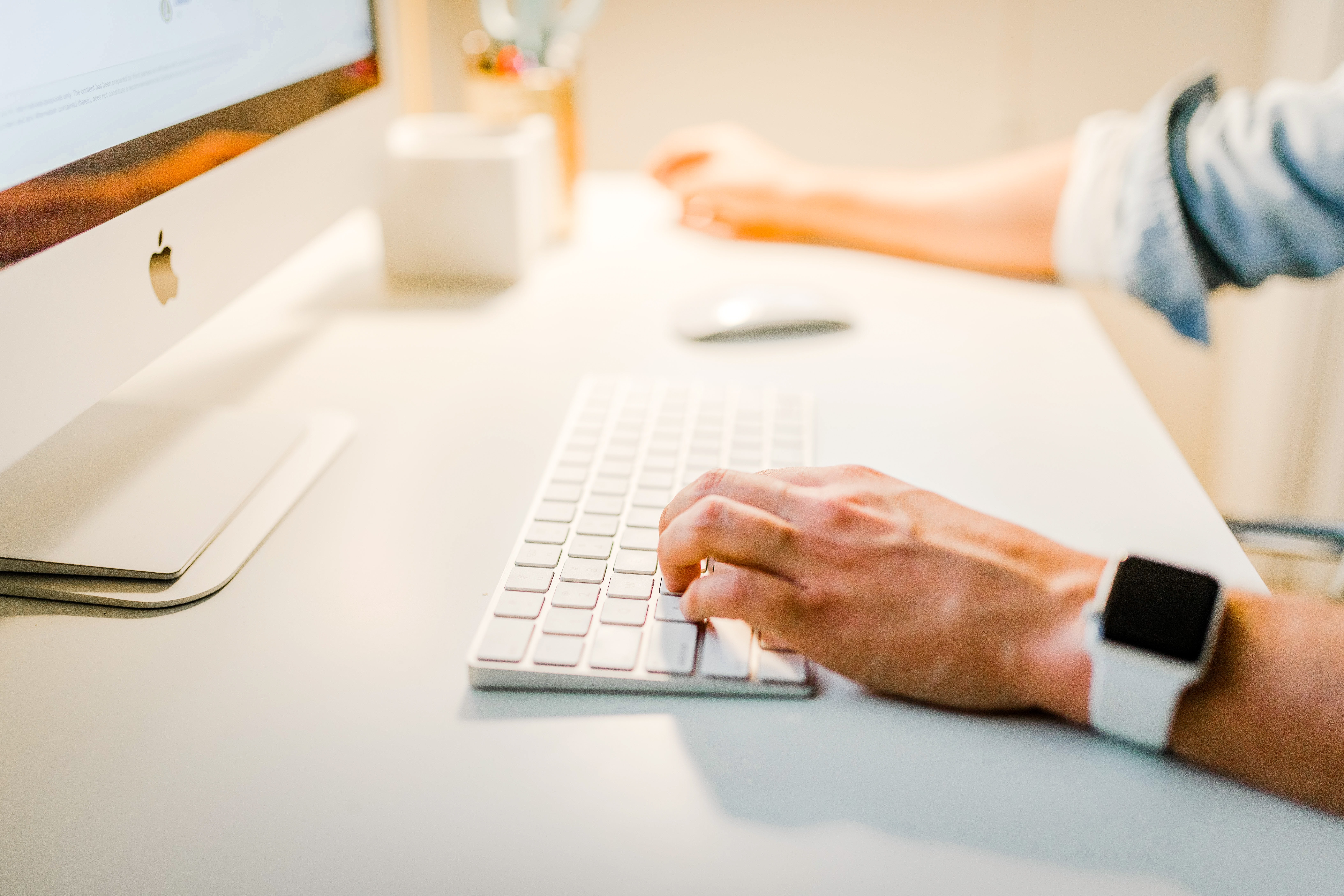 A person wearing a smartwatch is typing on a white keyboard in front of a computer monitor. Their other hand is on a mouse, and everything is set on a clean white desk.